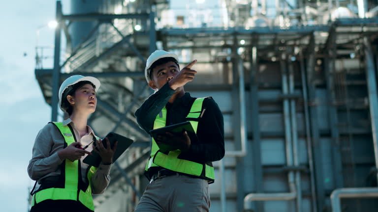 Two engineers wearing safety helmet and reflective vest using radio and tablet at construction site, coordinating inspection under blue sky, teamwork and professional project management concept.